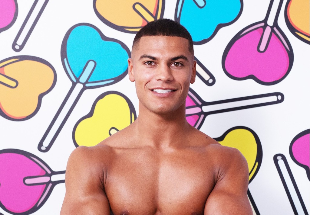A shirtless Reece Ford, a contestant on the Love Island 2022 series standing agains a backdrop of heart-shaped lollipops.